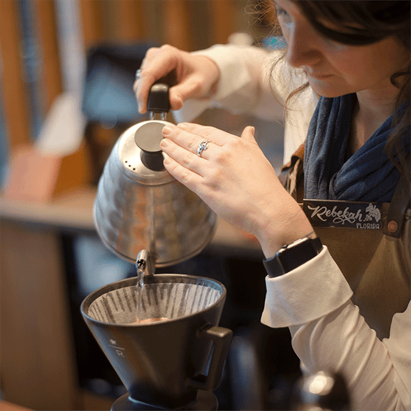 Woman with nametag "Rebekah" pouring water from a gooseneck kettle into a Starbucks Reserve-branded pour over
