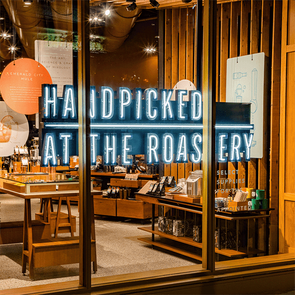 Storefront with neon sign that reads "Handpicked at the Roastery" with merchandise in the background