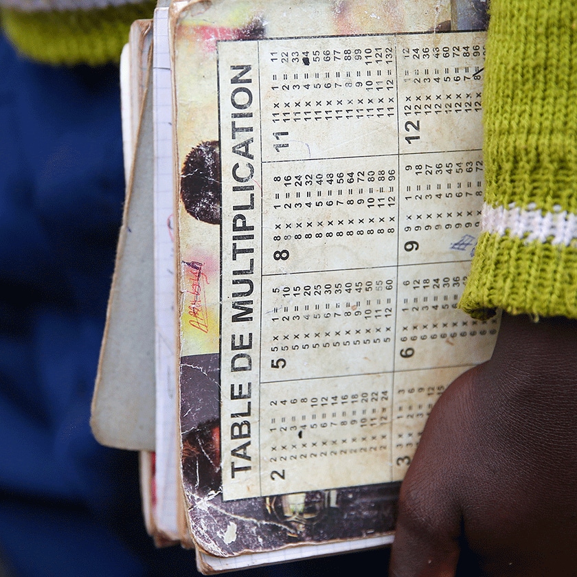 Closeup of a child's arm and hand holding a notebook that reads "Table de Multiplication"