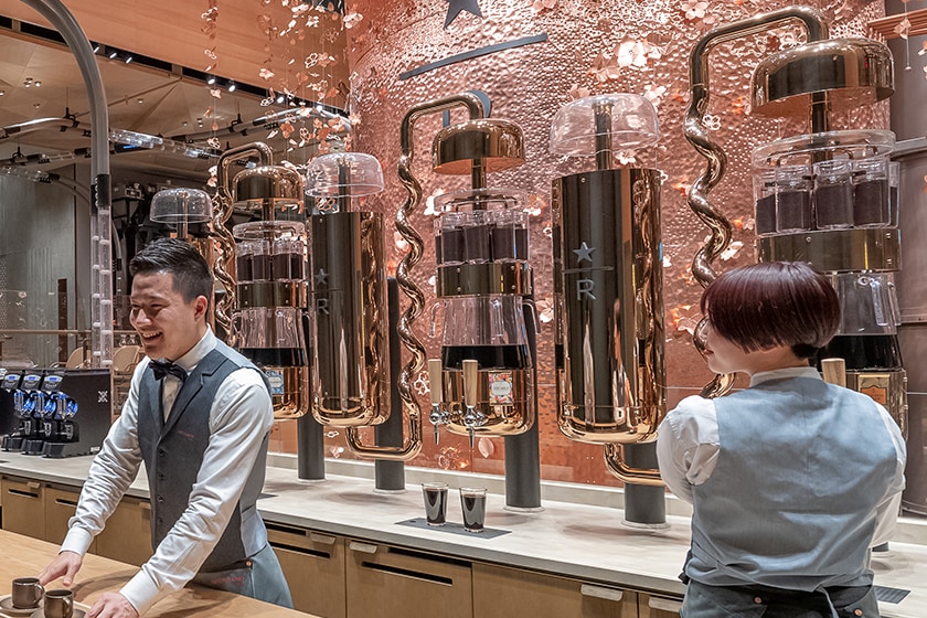 Two baristas in front of a row of cold brew silos which include intricate, curved pipes