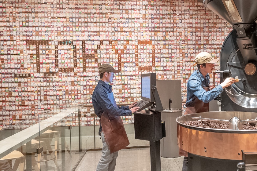 Display of hundreds of colorful Starbucks Reserve coffee cards on a large wall, with "TOKYO" spelled out within, and coffee roasting equipment in foreground