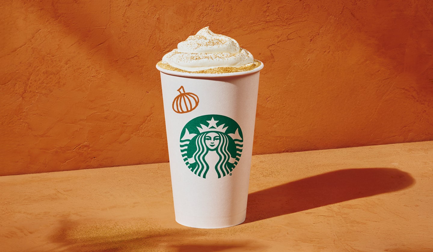 A Pumpkin Spice Latte in a hot to-go Starbucks® cup. An orange pumpkin is handdrawn on cup. It sits in front of an orange backdrop.