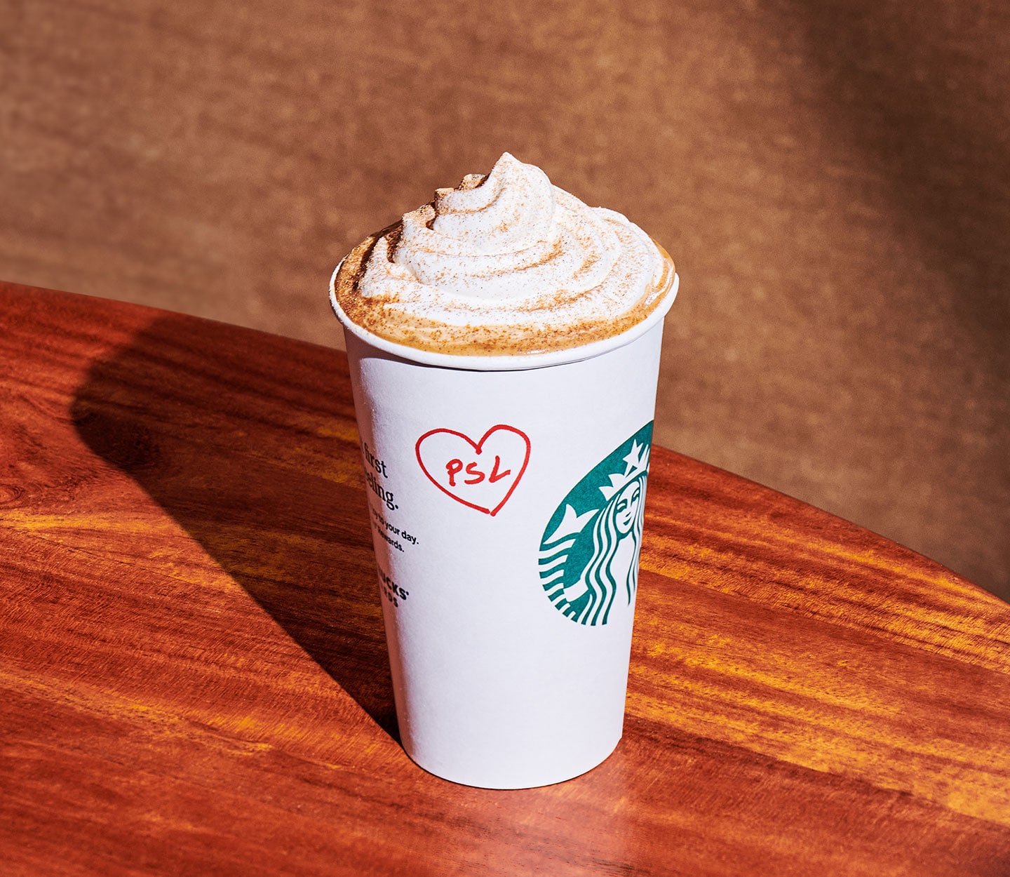 A hot coffee drink topped with whipped cream in a white Starbucks cup rests on a wooden countertop. On the side of the cup, in red pen are the letters, “PSL,” inside of a hand drawn heart.