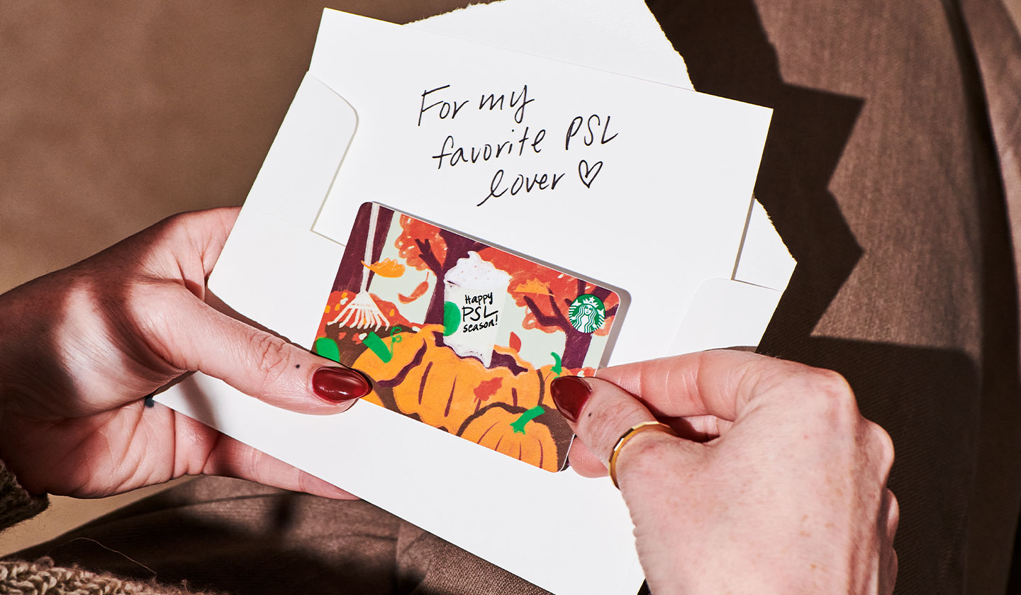 A seated person holds an opened envelope and a colorful fall-themed Starbucks® gift card in their hands. An accompanying note includes the message, “For my favorite PSL lover,” with a drawing of a heart.