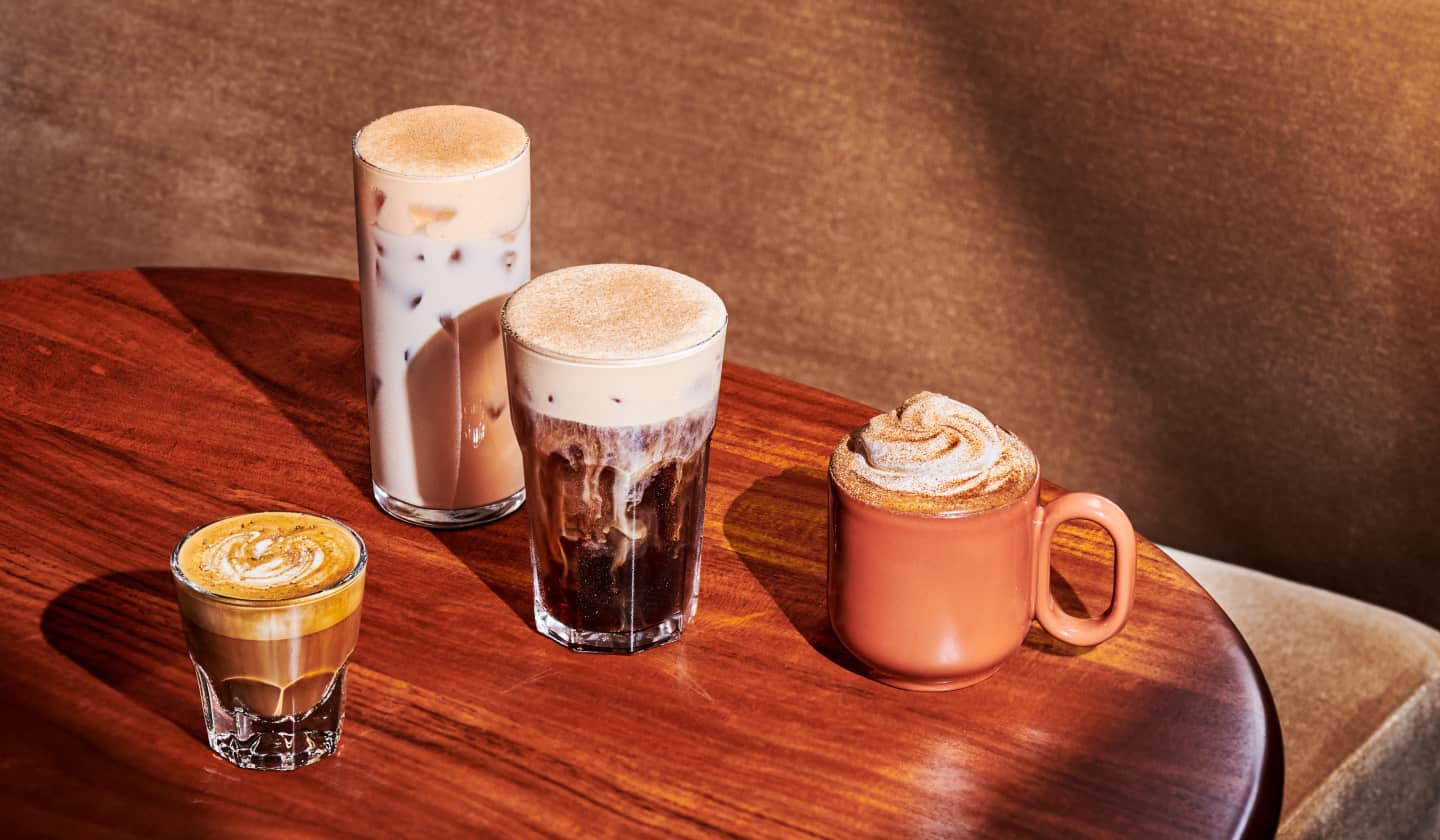 Four fall drinks in mugs and glasses on a wood surface in front of a brown textured background.