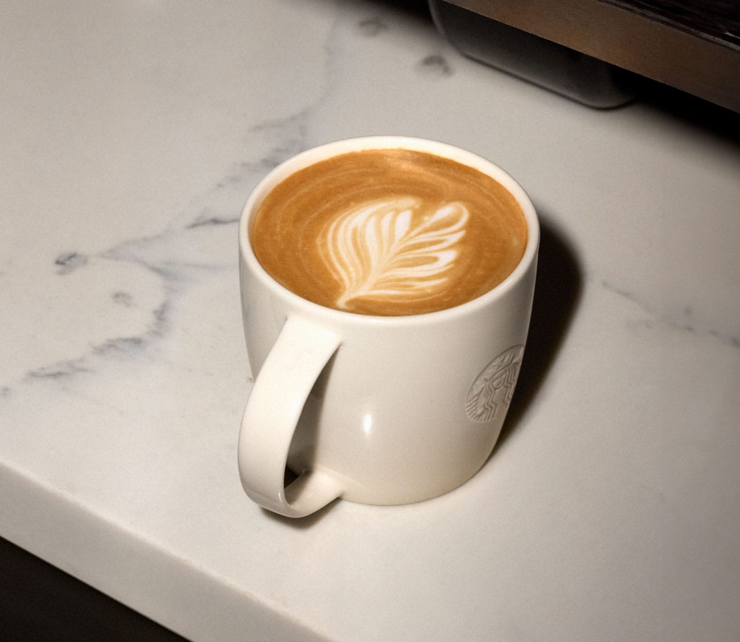 A hot coffee drink with a foam and leaf art in a white mug is resting on a white marble countertop.