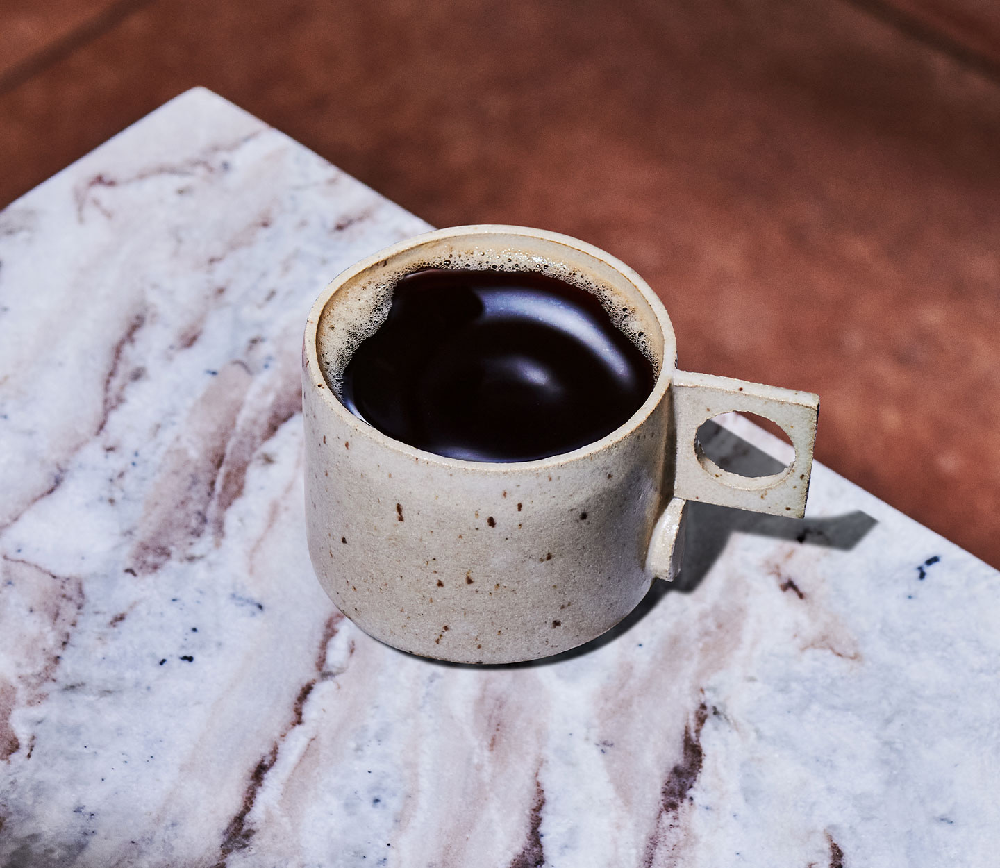 Hot coffee in a speckled off-white mug sits on a veined white marble countertop.