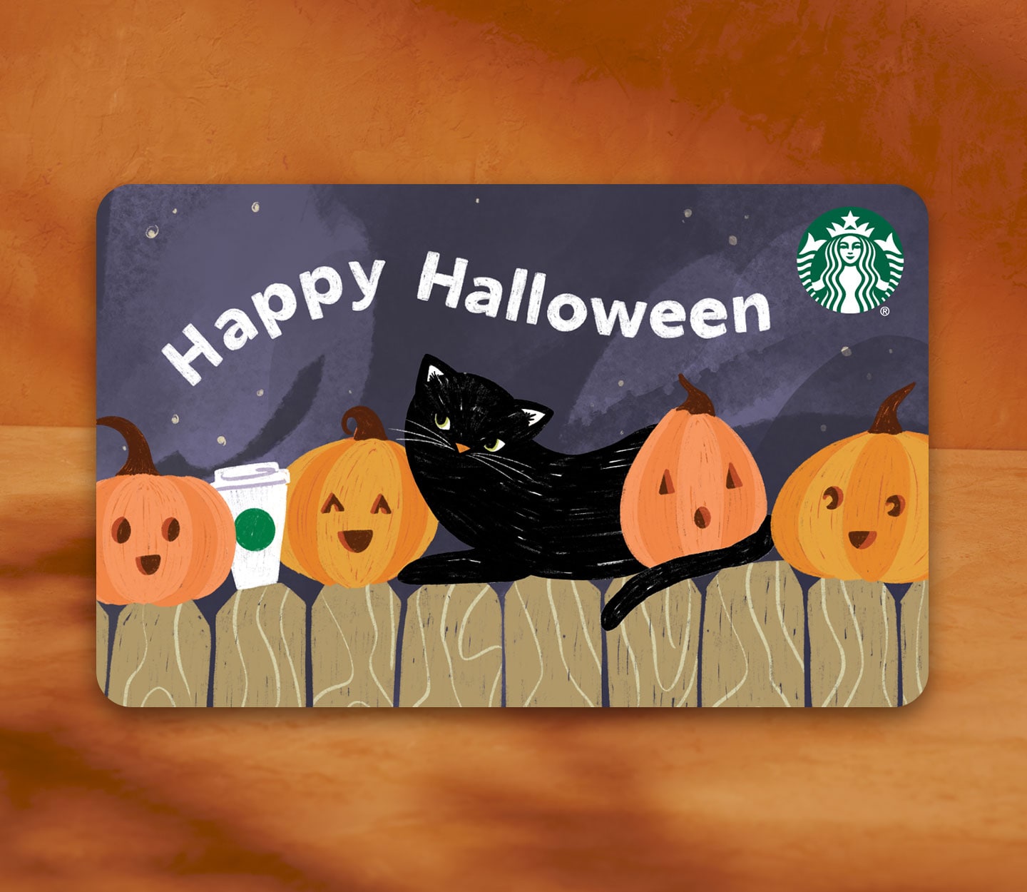 A Starbucks gift card with the words, “Happy Halloween” in white over an illustration of a dark sky and a brown fence topped with a row of orange pumpkins, a Starbucks cup and black cat.