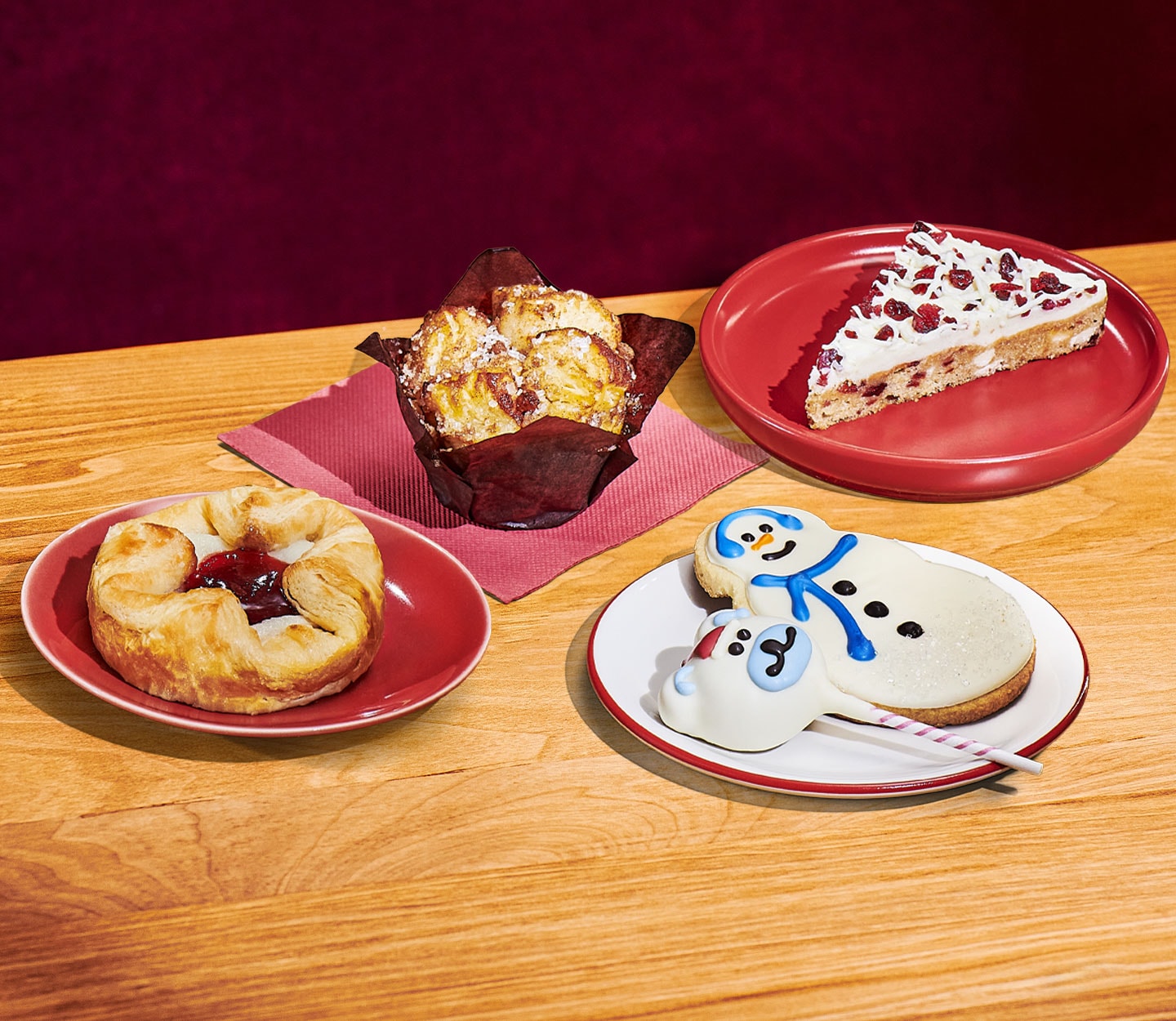 Five bakery items: a round pastry with dark red filling on a red plate; a flaky pastry with sprinkles in a brown wrapping on a red napkin; a triangular cake slice with white frosting and red berries on a red plate; and a white frosted cookie with snowman design next to a round cake on a stick with a white polar bear head design.
