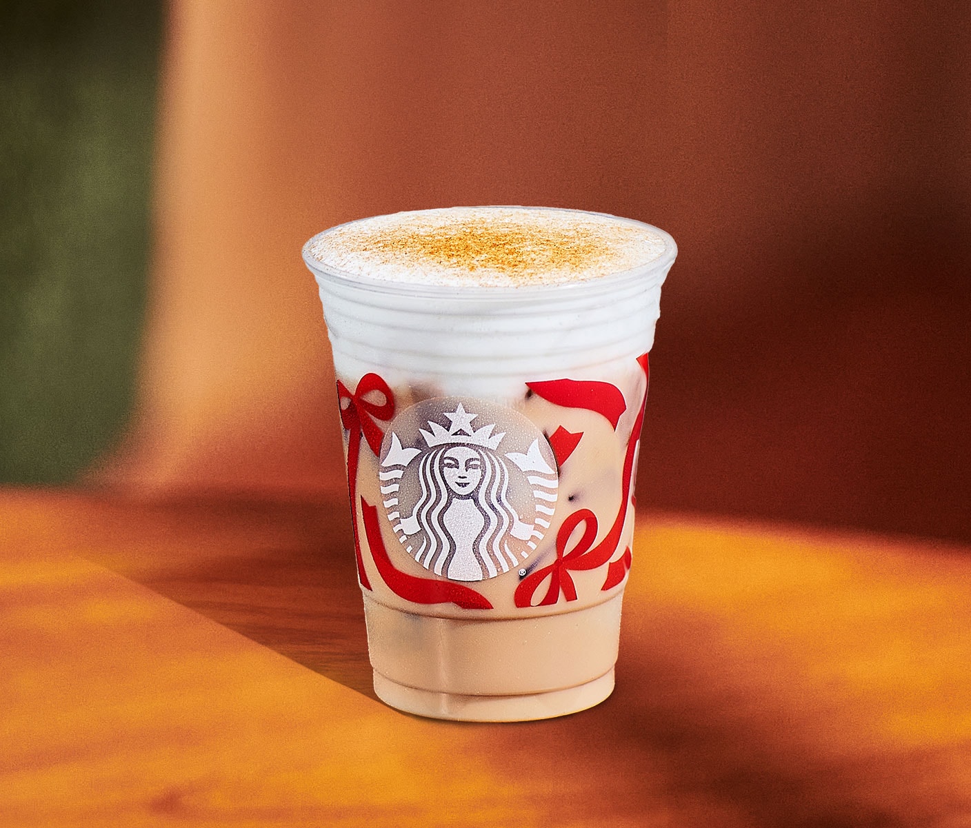 A creamy iced tea drink with foam top and brown spice toppin in a clear Starbucks cup with a red ribbon design.