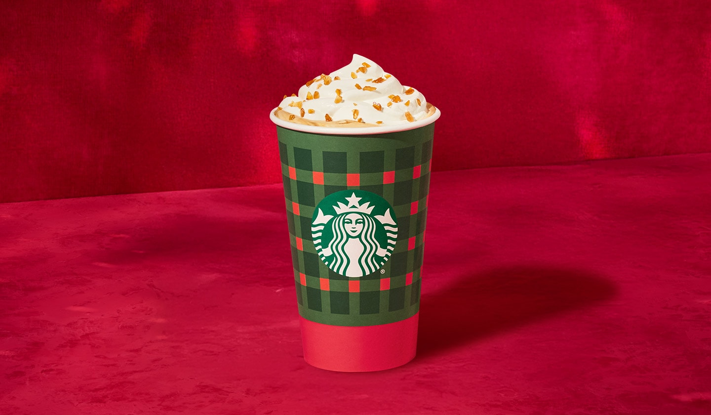 A Caramel Brulée Latte in a holiday hot to go cup set against a red velvet backdrop.