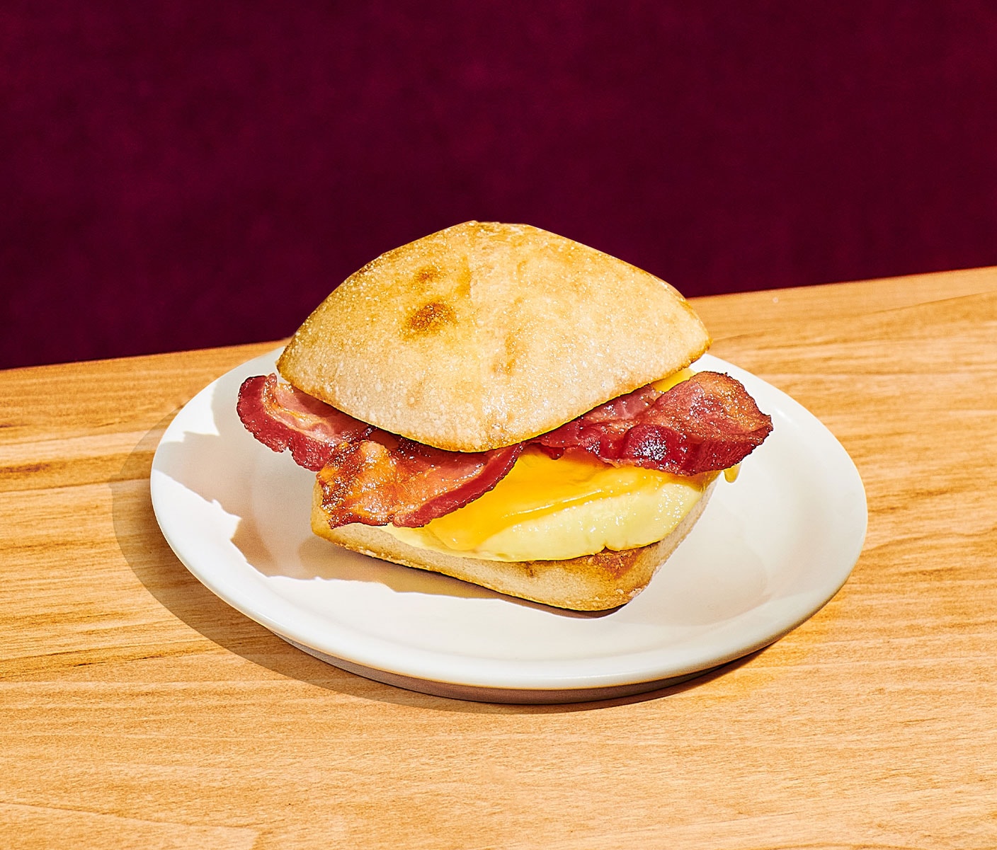 A breakfast sandwich with layered egg, cheese and bacon between a roll on a white plate.