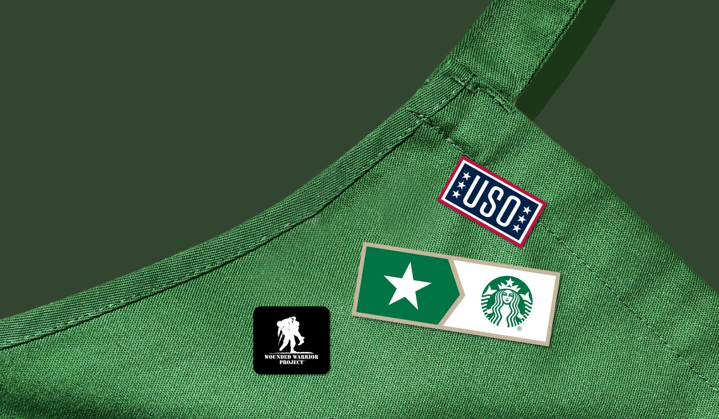 Close-up of a Starbucks green apron, decorated with a Wounded Warrior Project badge, a USO pin, and a Starbucks veteran pin.