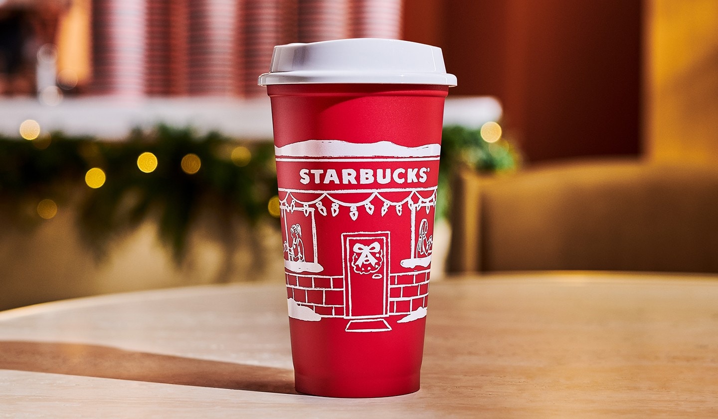 A Starbucks reusable red cup with a festive storefront illustration rests on a wood surface.