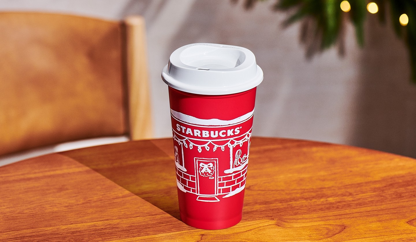 A Starbucks reusable red cup with a festive storefront illustration rests at the center of a wood table.