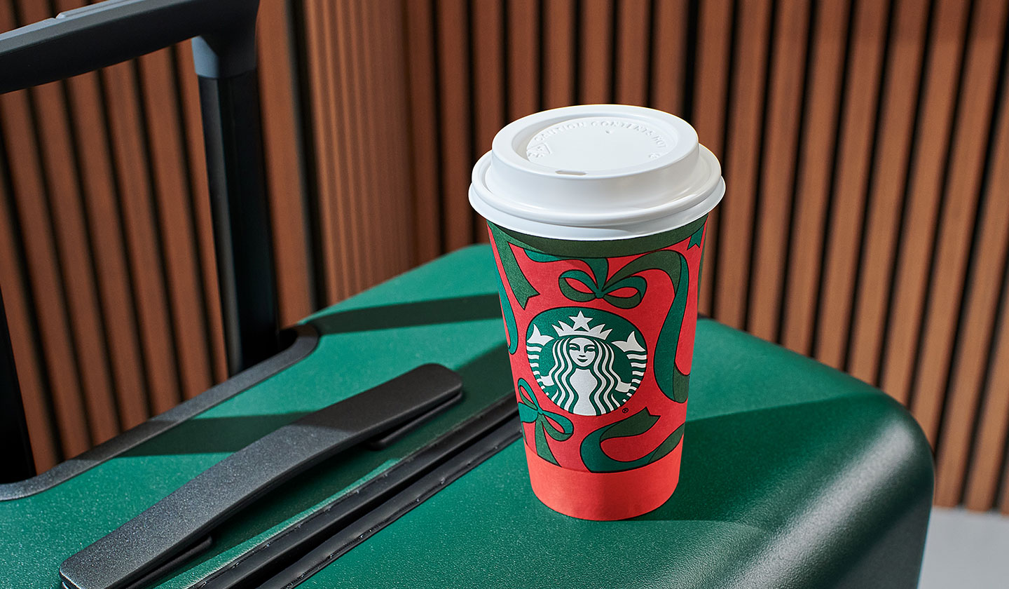 A Starbucks holiday cup with a red and green giftwrap design sits on top of a green hardshell suitcase.