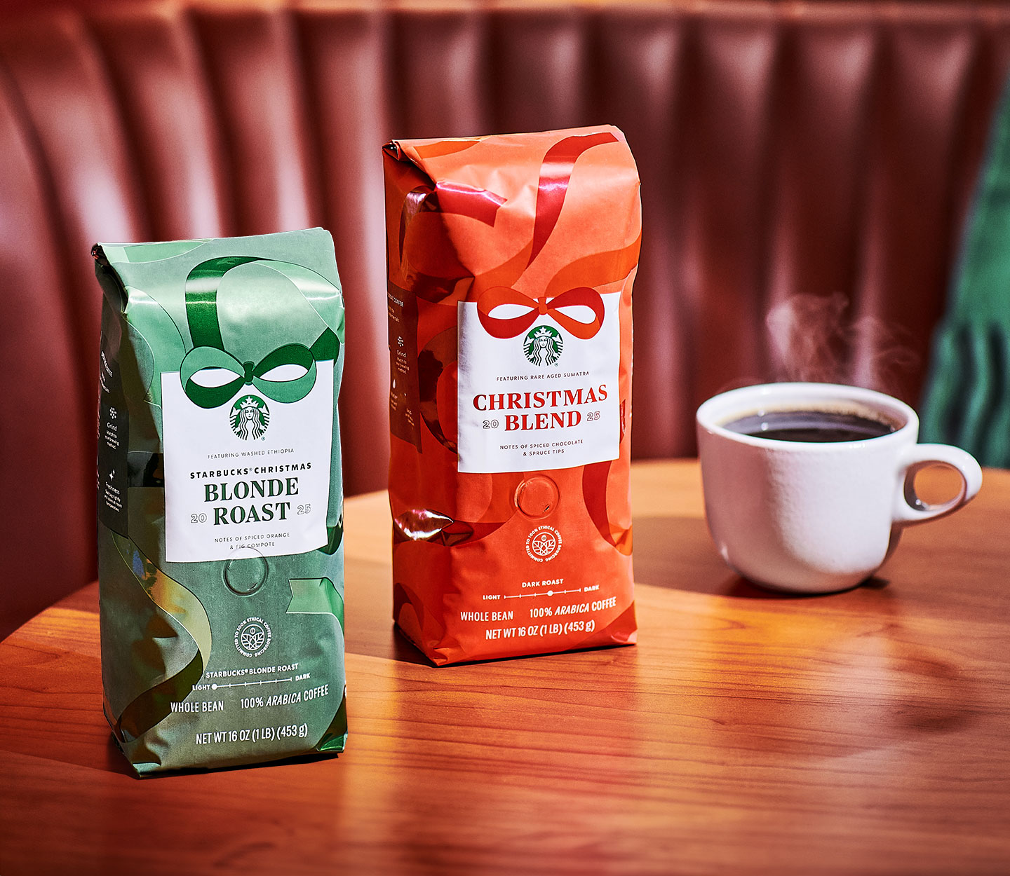A white mug of hot coffee on a table next to two bags of coffee with ribbon designs. One is green with the words, “Starbucks® Christmas Blonde Roast”; the other is red with the words ”Starbucks® Christmas Blend.”