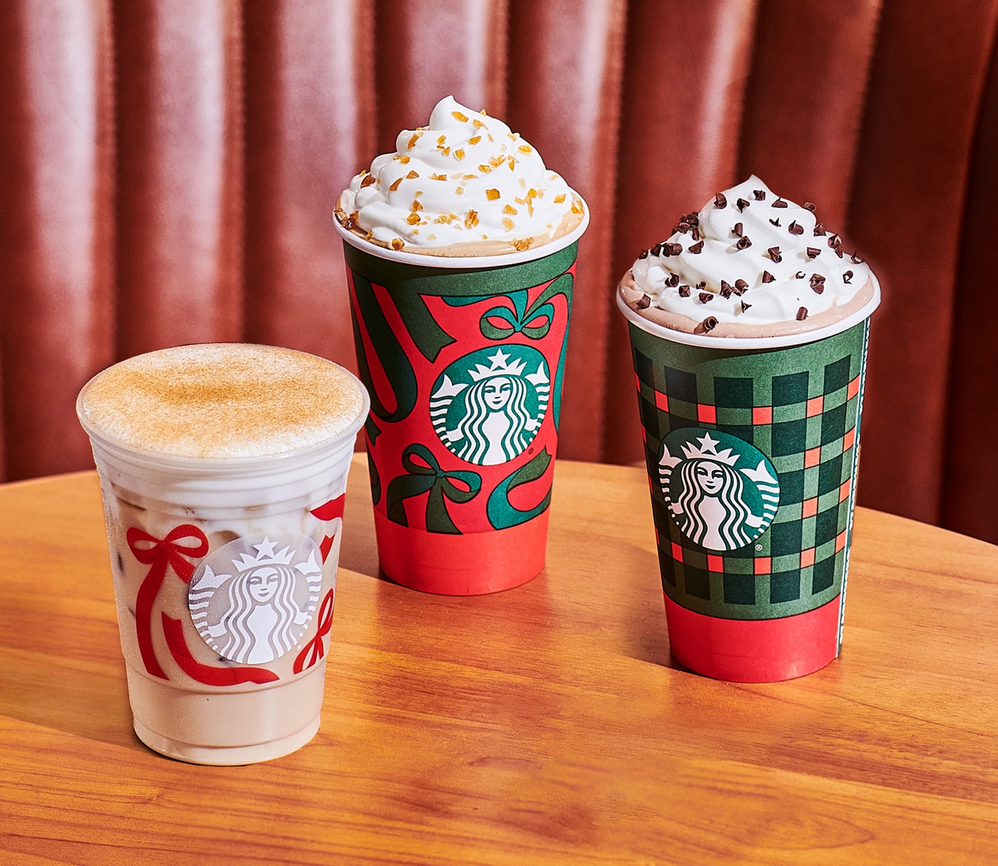 Three Starbucks holiday drinks in festive red and green to-go cups sit side by side on a wooden table in front of a pleated brown leather booth.