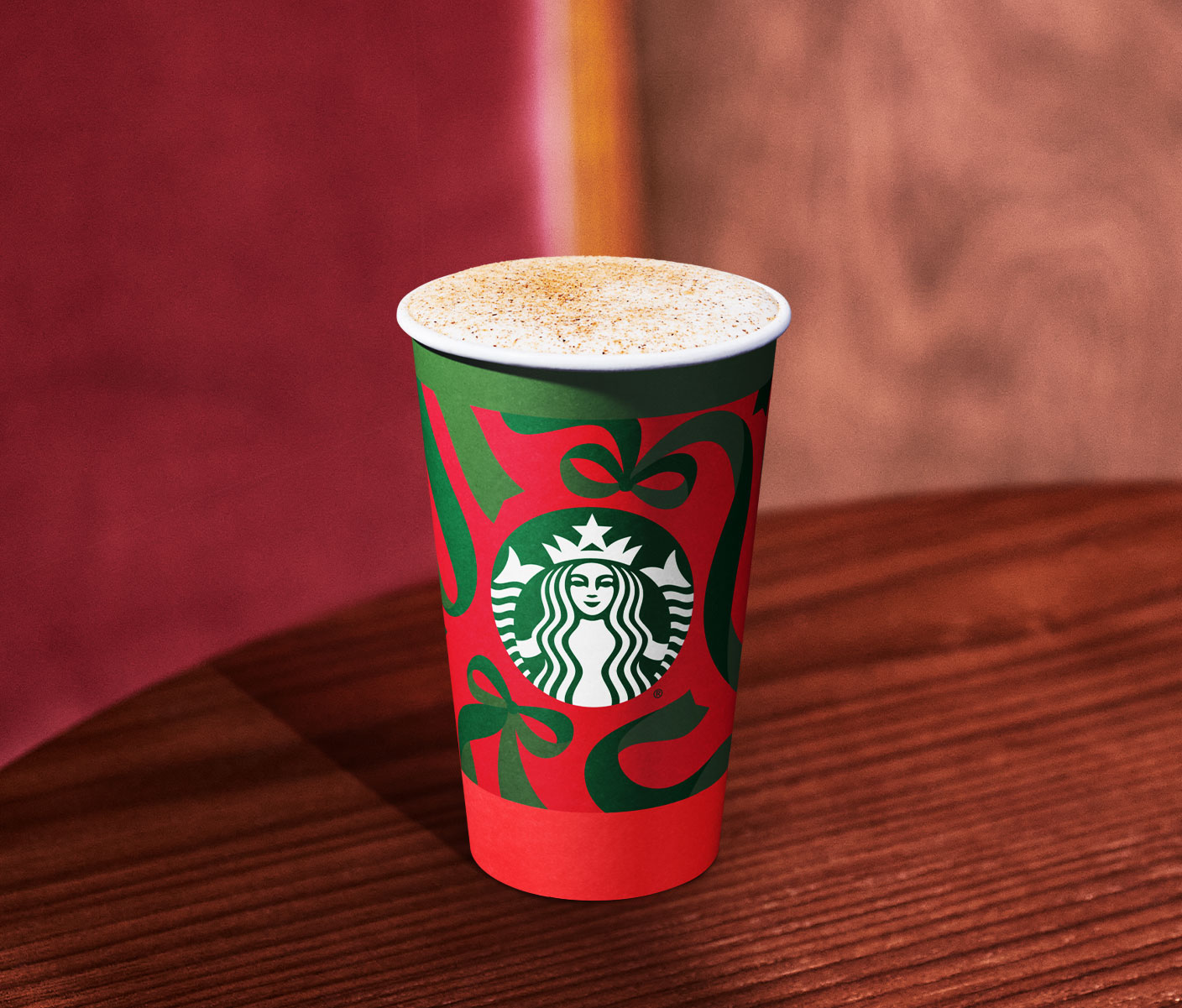 A hot drink with a foamy top and brown sprinkles in a red cup with green ribbons and the Starbucks logo, placed on a wood table.