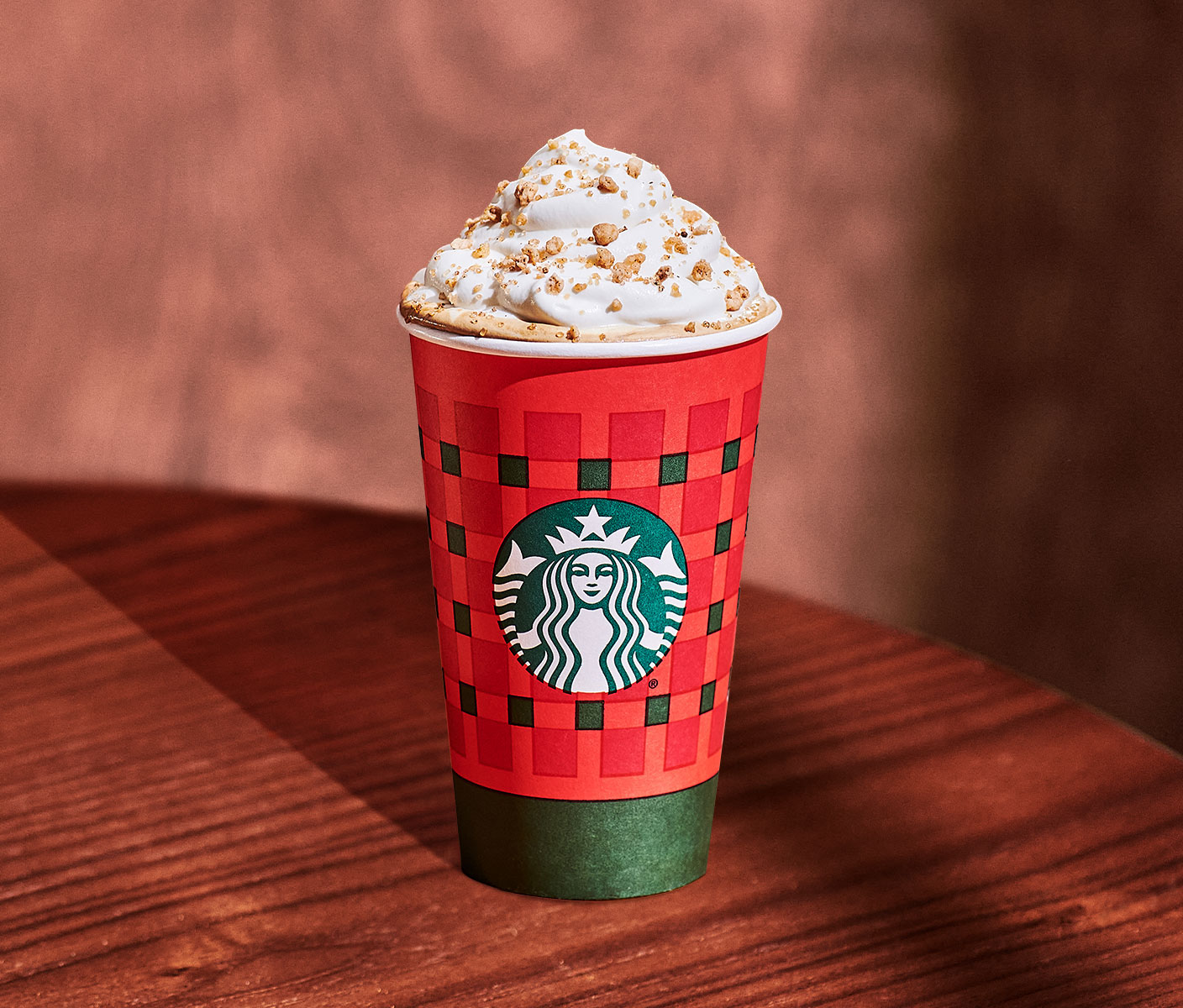 A hot drink topped with whipped cream and brown sprinkles in a red cup with green ribbons and the Starbucks logo, placed on a wood table.