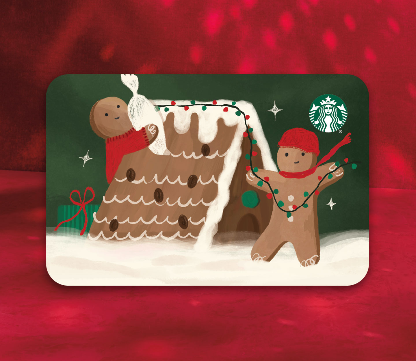 A Starbucks gift card with two gingerbread figures decorating a gingerbread house with frosting and lights, in the snow. One is wearing a red hat and scarf, the other a red sweater.