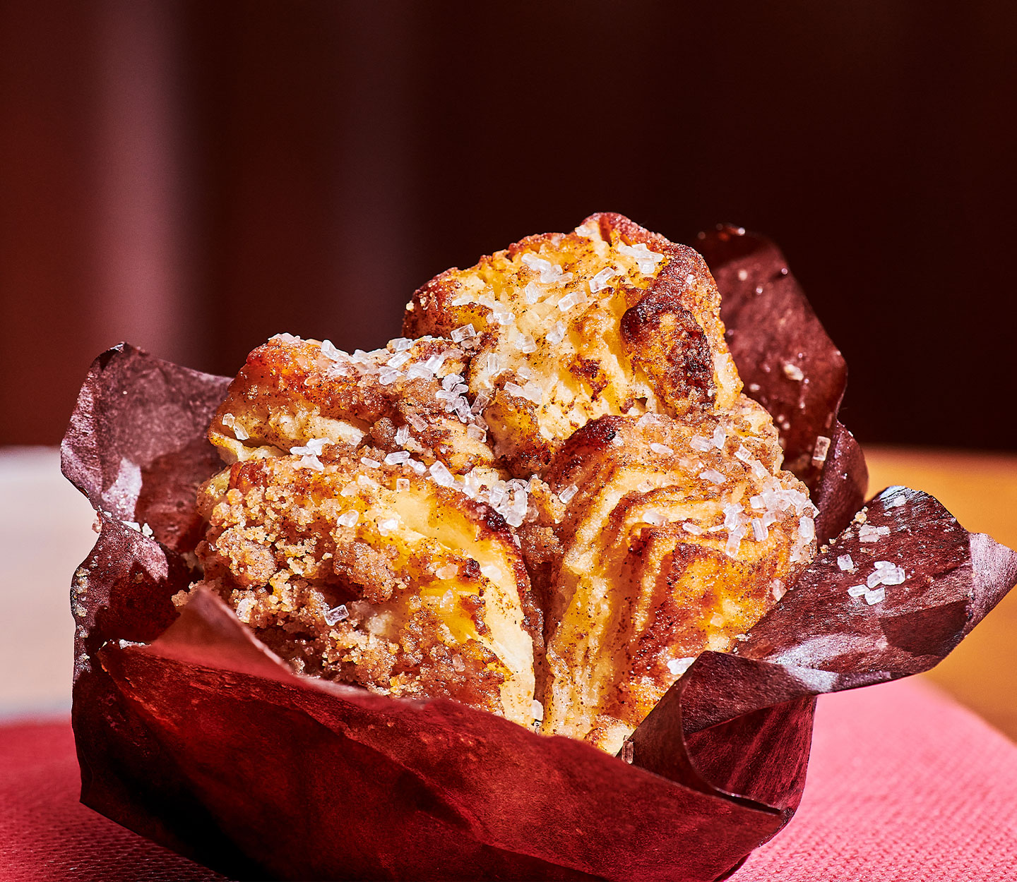 The top of a golden flaky pastry with white sprinkles on top in a burgundy paper wrapper.