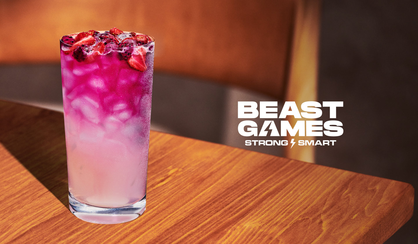Close-up shot of an iced lemonade drink with fruit inclusions on a wooden surface. Next to it is the Beast Games Season 2 Strong versus Smart show logo. 