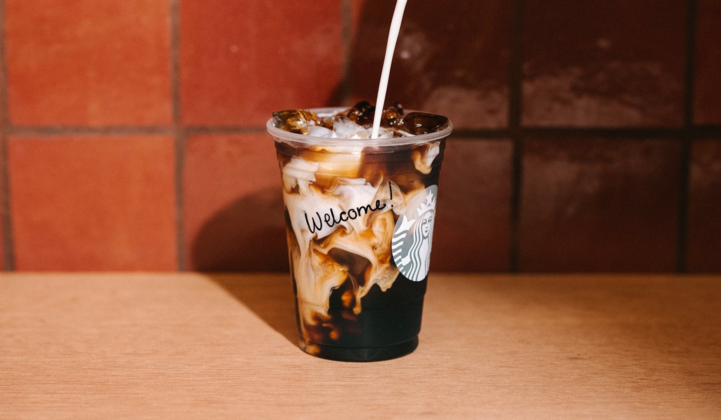 A Vanilla Sweet Cream Cold Brew in a to-go cup with a handwritten "welcome" message and a stream of cream being poured in the cup.