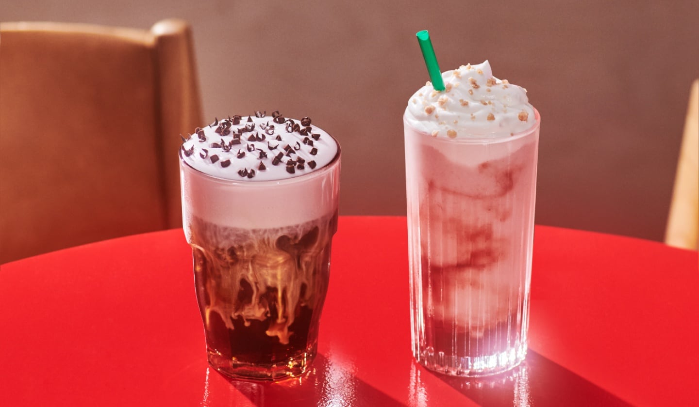 A White Chocolate Strawberry Cream Cold Brew and Strawberry Shortcake Frappuccino® blended beverage sit next to each other on a red table.