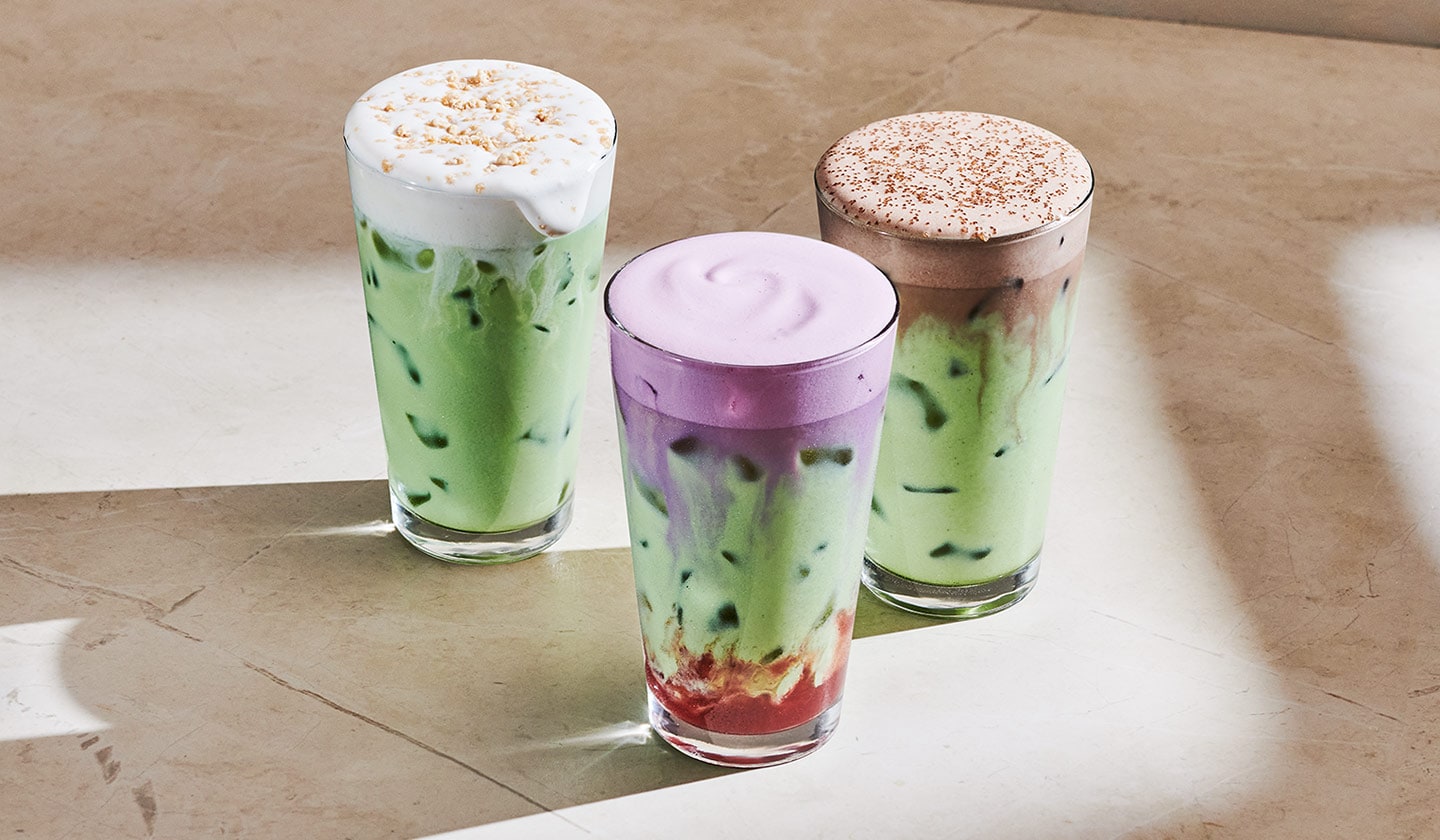 Three colorful iced matcha drinks topped with cold foam in clear glasses on a textured beige background.
