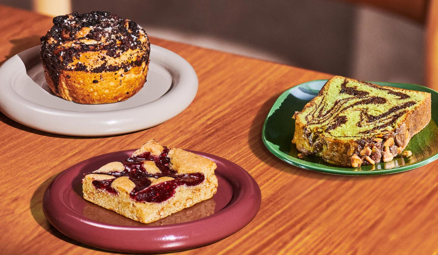 Three bakery items on individual plates: a Cookie Croissant Swirl on the left, a Berry Blondie bar in the center and a slice of Chocolate Pistachio Loaf on the right.