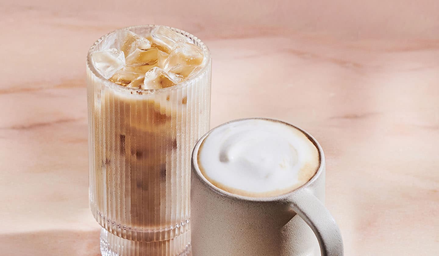 An iced chai drink in a tall glass and a hot chai drink with foam in a mug rest on a lightly marbled surface.
