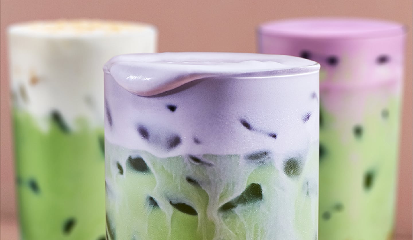A trio of layered matcha drinks stand tight in the foreground, with a focus on the colorful cold foam at their surface.