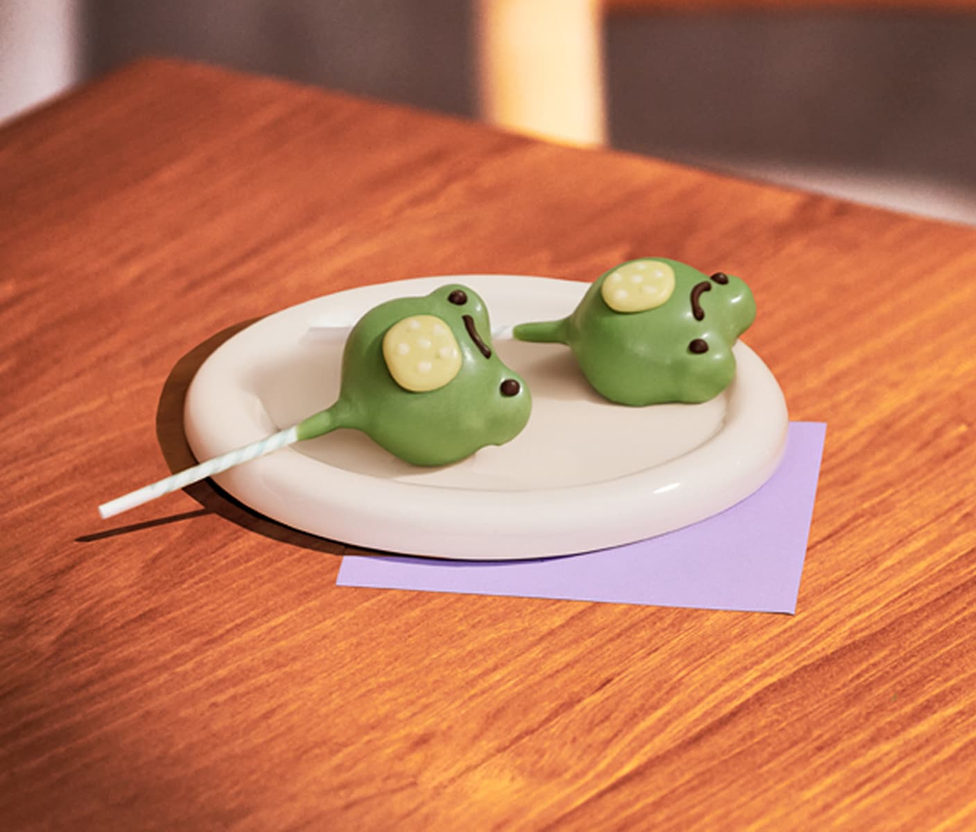 Two roundish cake balls on sticks, frosted with a smiling frog face design
