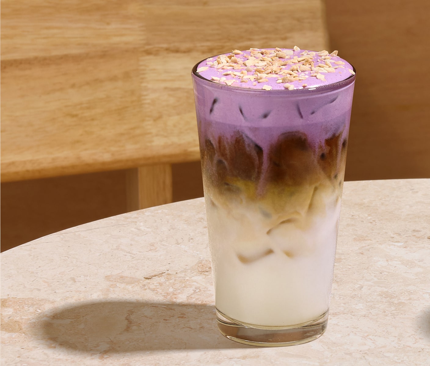Iced espresso with milk in a clear glass, finished with pastel purple cold foam and toasted coconut flakes, placed on a marbled beige surface in front of a wood background.