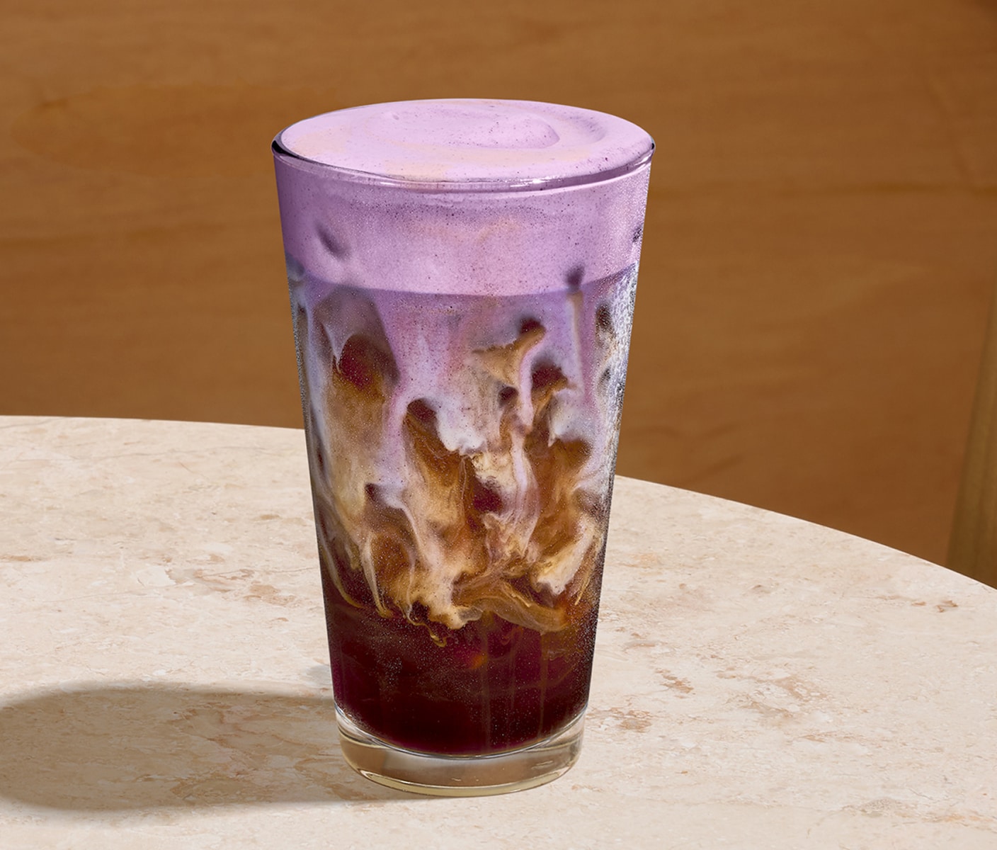 Iced espresso with swirls of milk in a clear glass, finished with cascading purple cold foam, placed on a marbled beige surface in front of a wood background.