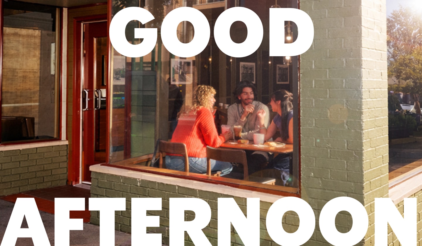Exterior view of a Starbucks with three people visible through the window, chatting at a table with drinks and snacks. Text on the image reads "Good afternoon."