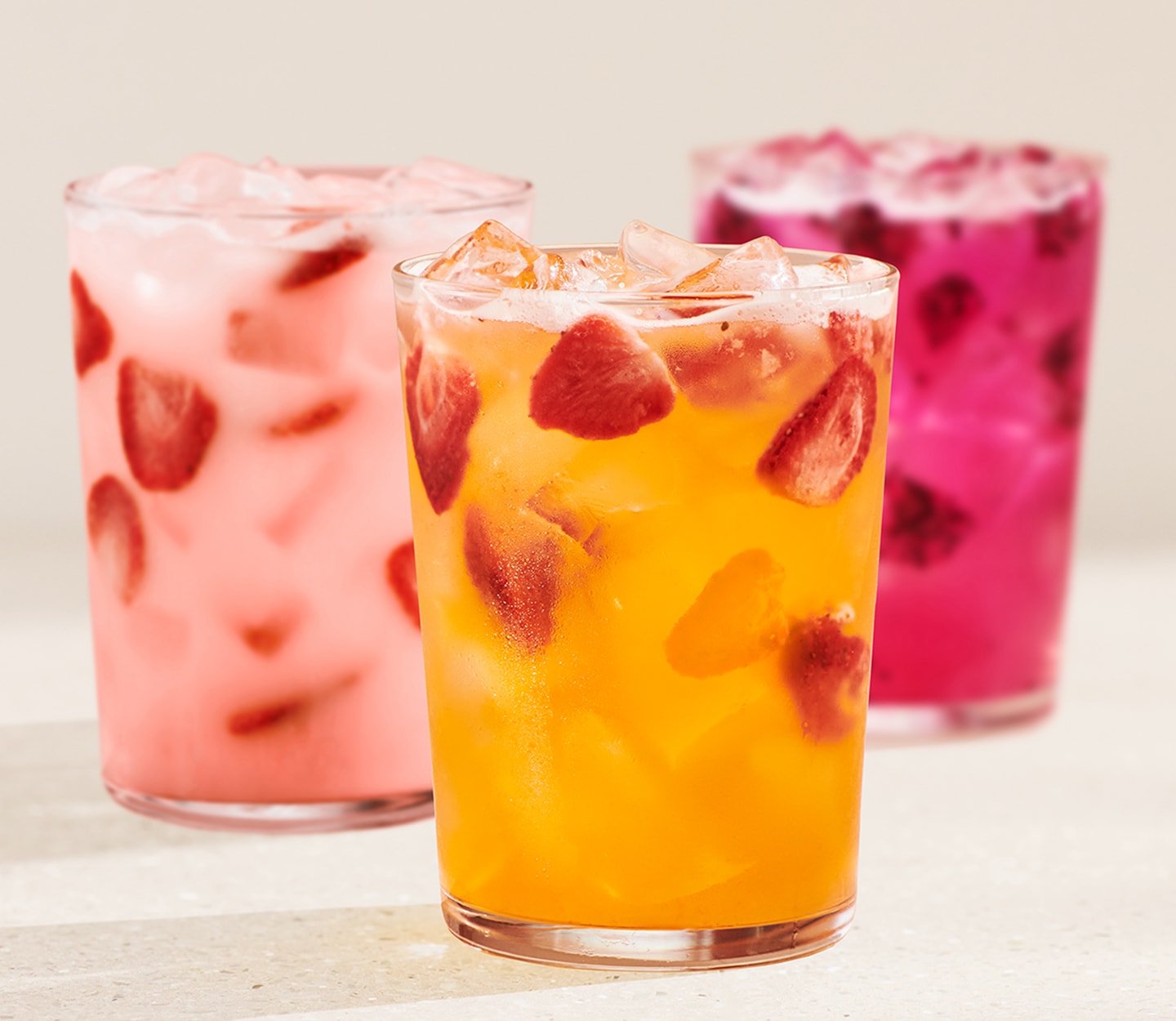 A Pink Energy Drink, a Mango Strawberry Energy Refresher and a Mango Dragonfruit Energy Refresher in tall glasses rest on a speckled surface.