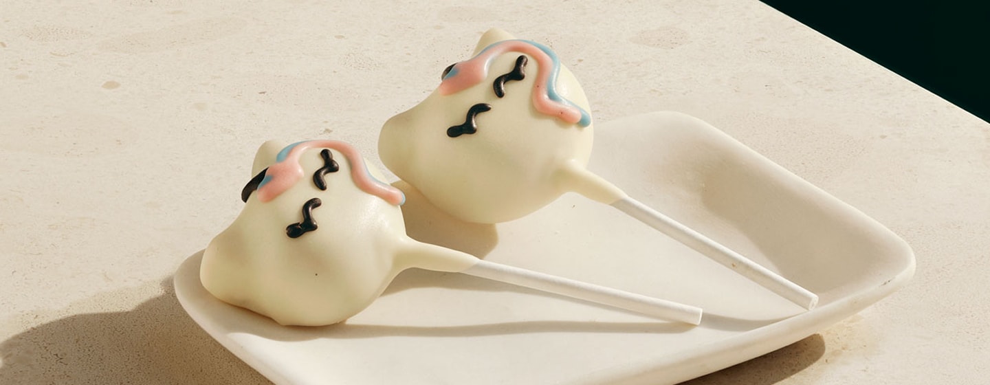 Two small roundish cake pops with unicorn face design on sticks on a plate.