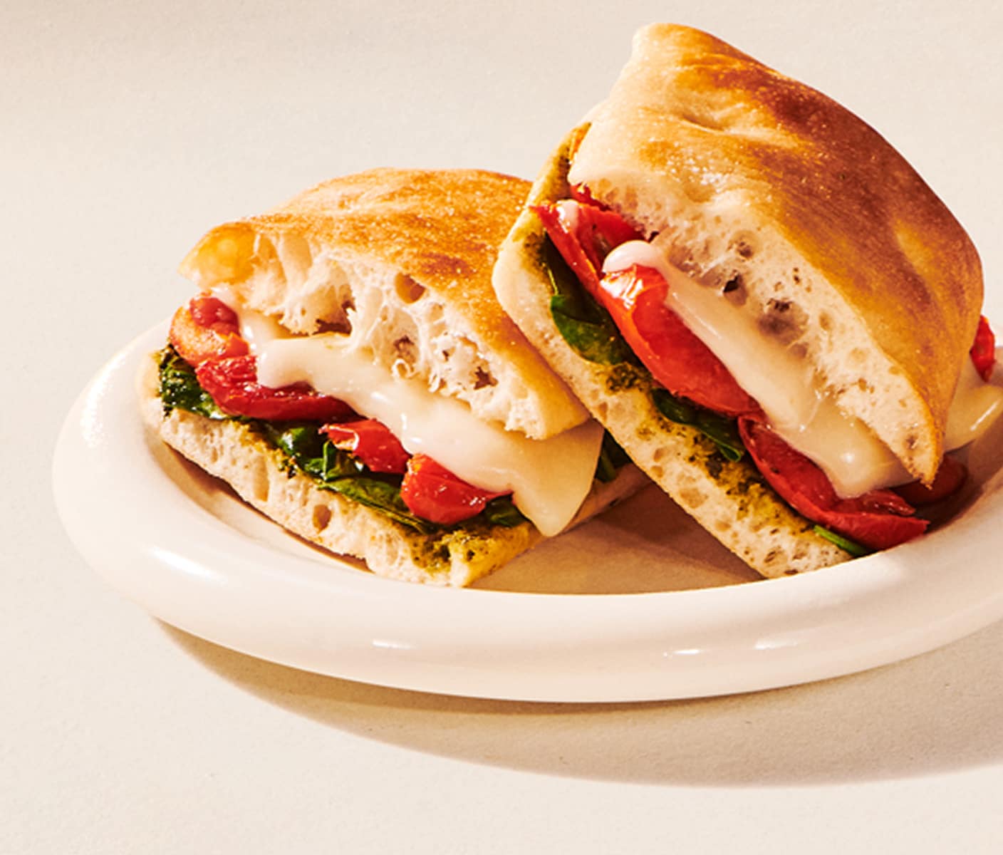 Two halves of a toasted focaccia sandwich filled with spinach, pesto, cheese and tomato are stacked on a plate.