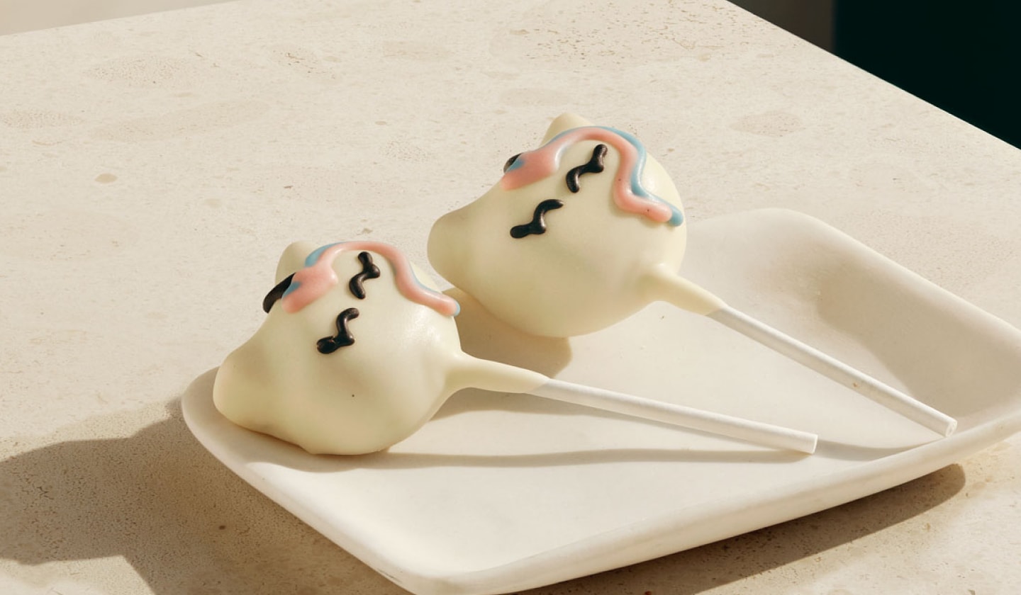 Two small roundish cake pops with unicorn face design on sticks on a plate.
