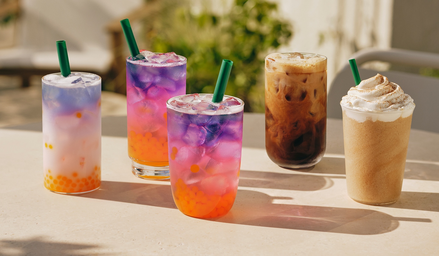 Three pink, purple and orange layered drinks, an iced coffee with milk and a blended drink with whipped cream in glassware on a sun-drenched table outside.