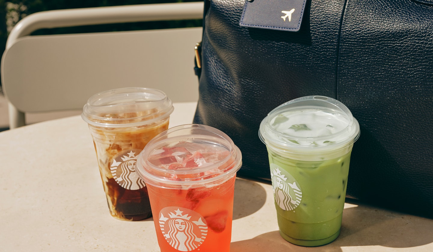 An iced coffee drink, a Refresher and an iced matcha latte on a countertop next to a weekender bag with a travel tag on the handle.