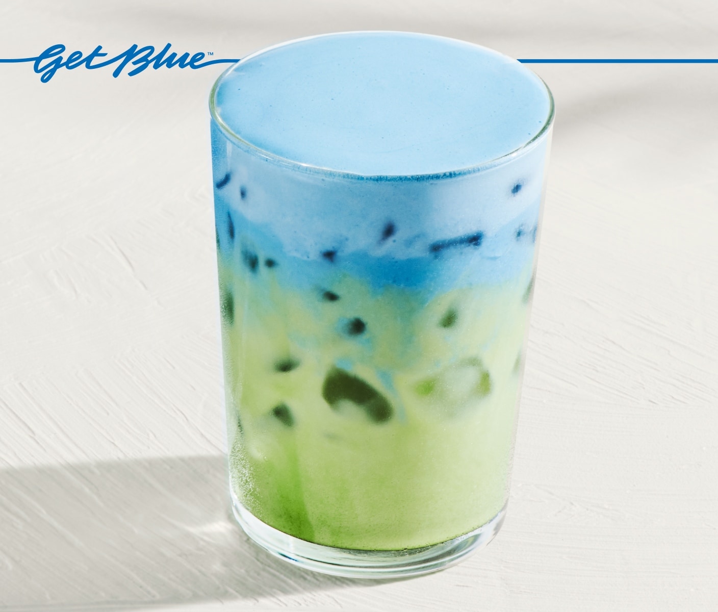 An iced matcha with light-blue cold foam in glassware on a light-gray surface. Blue script that reads “Get Blue” sits above it.