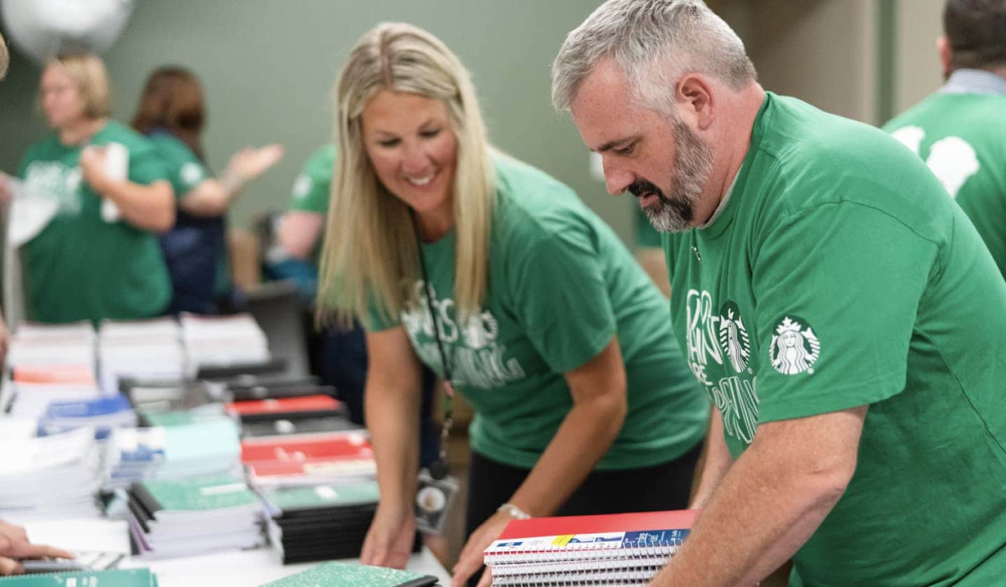 A woman and man wearing green shirts with the Starbucks logo on the sleeve work at a table stacked with notebooks
