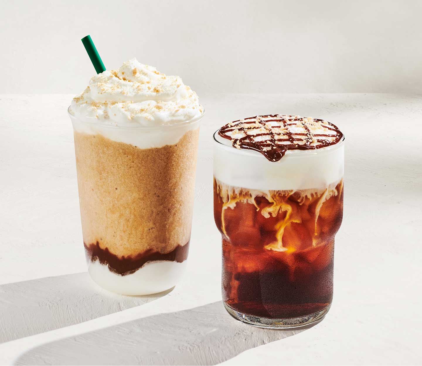 A blended coffee drink with chocolate sauce and whipped cream next to an iced coffee drink with white cold foam and chocolate drizzle, against a light-gray background.
