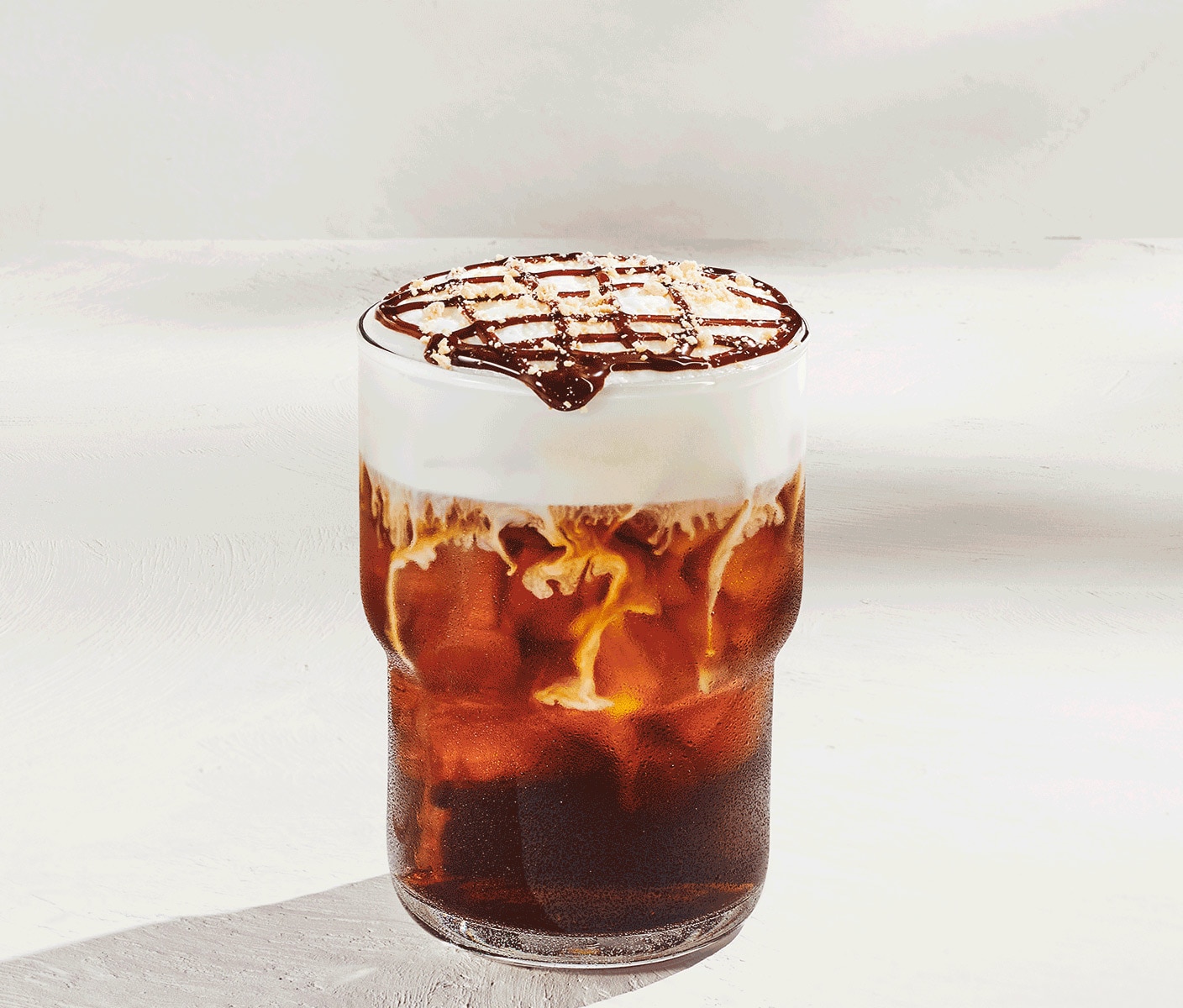 An iced coffee drink with white cold foam, chocolate drizzle and graham-cracker crumble against a light-gray background.