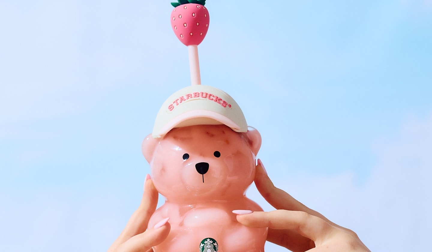 Two hands holding up a Starbucks Bearista cold cup in a white cap with a strawberry-topped straw.