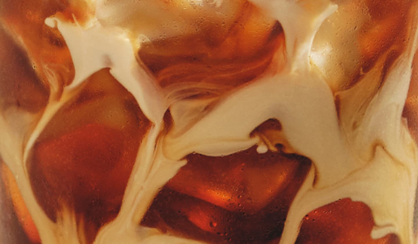 An extreme close-up of an iced coffee drink with coffee and cream swirling around chunks of ice through the glass.