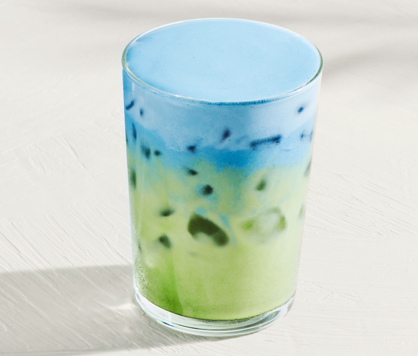An iced matcha with light-blue cold foam in glassware on a light-gray surface.